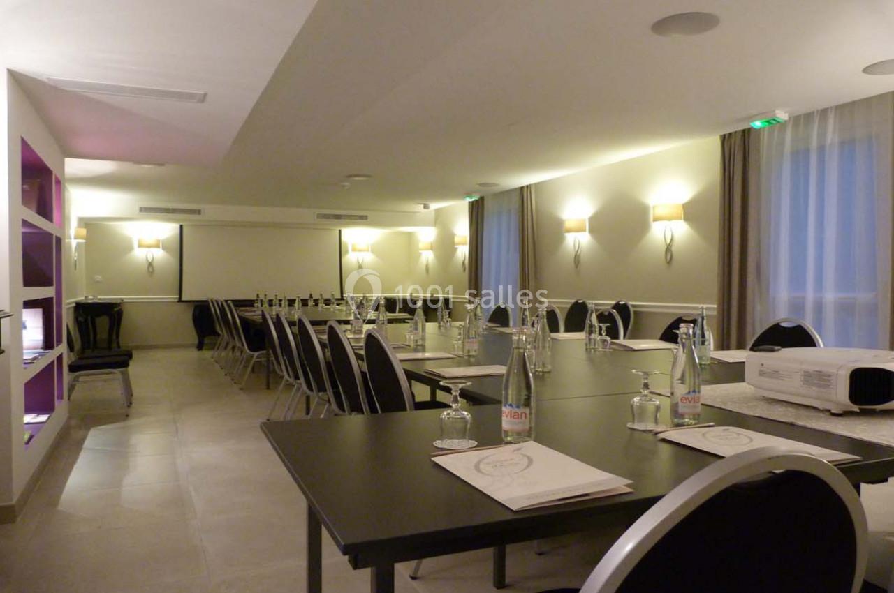 Salle de réunion lumineuse avec tables noires disposées en U, bouteilles d'eau, documents et vidéoprojecteur.