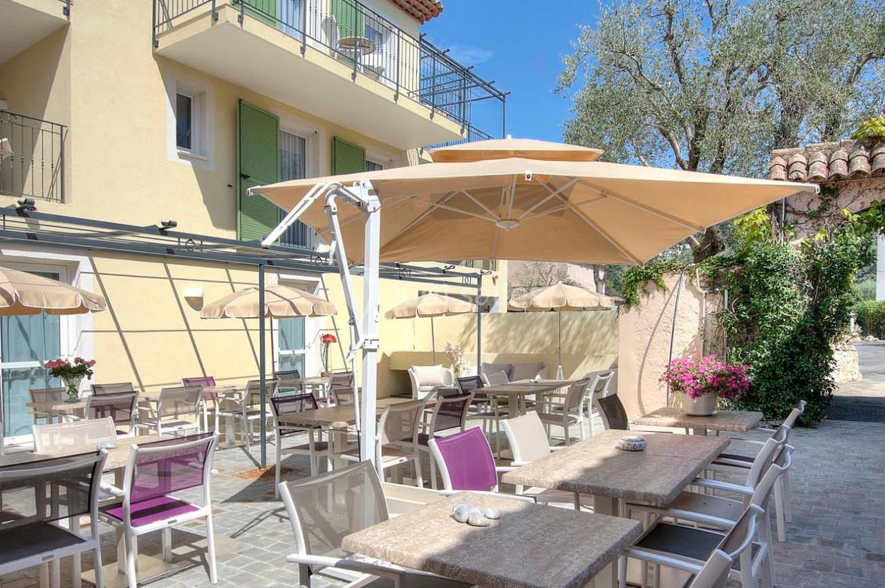 Terrasse extérieure avec tables, chaises et parasols, située devant un bâtiment beige à volets verts.