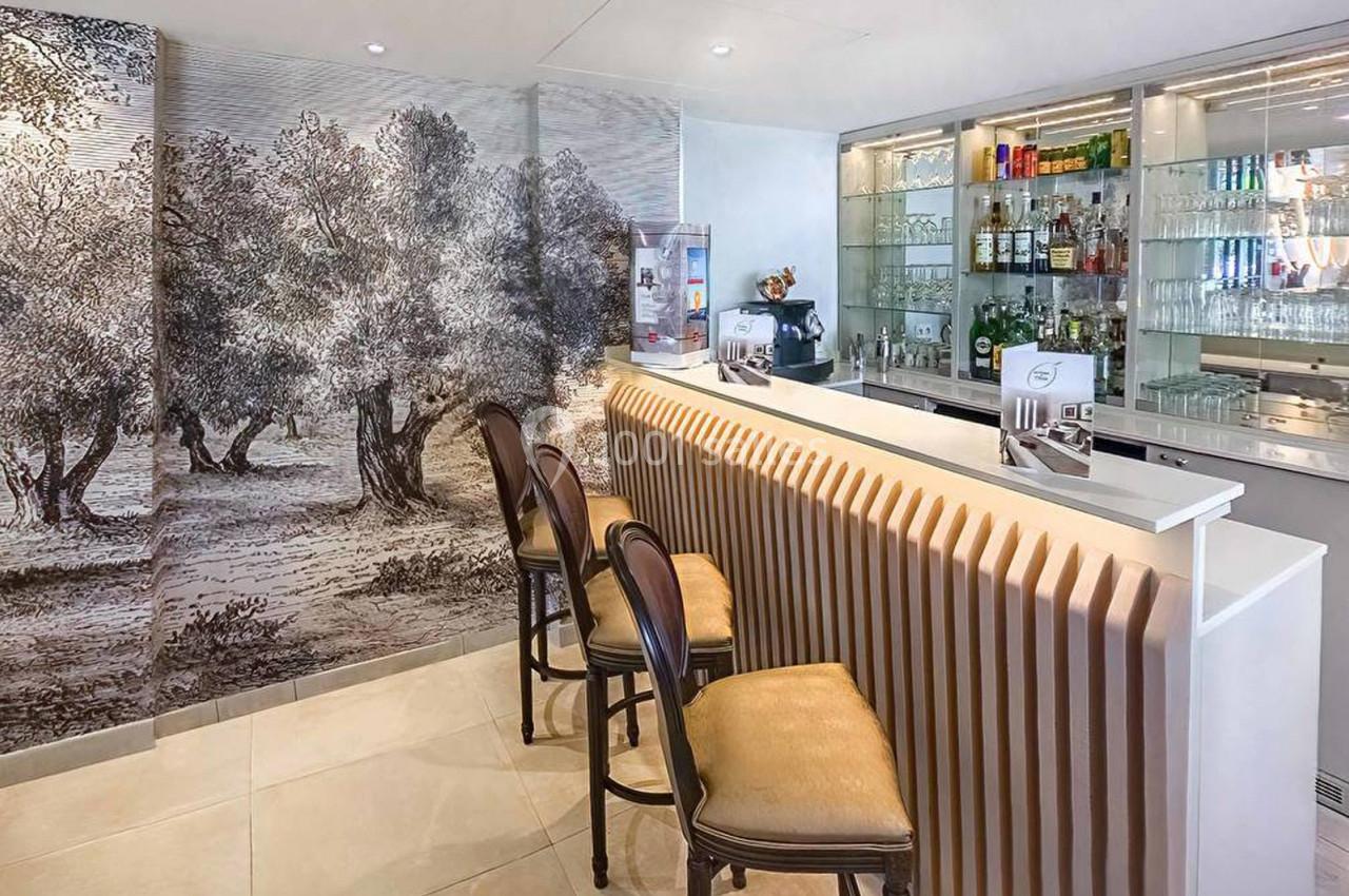 Bar moderne avec comptoir ondulé, trois tabourets et mur décoré d'une fresque représentant un paysage d'oliviers.