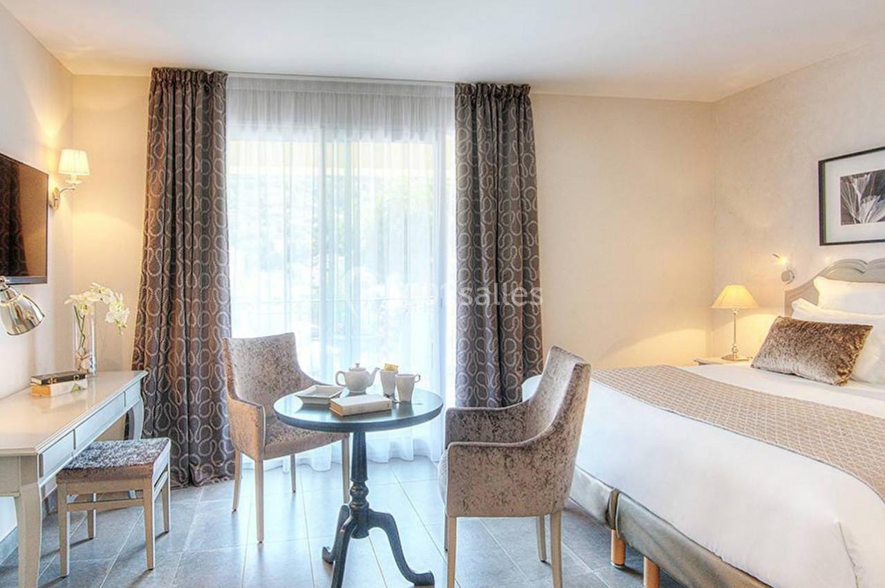 Chambre lumineuse avec lit double, table ronde, deux fauteuils, bureau, télévision murale et grande fenêtre avec rideaux.