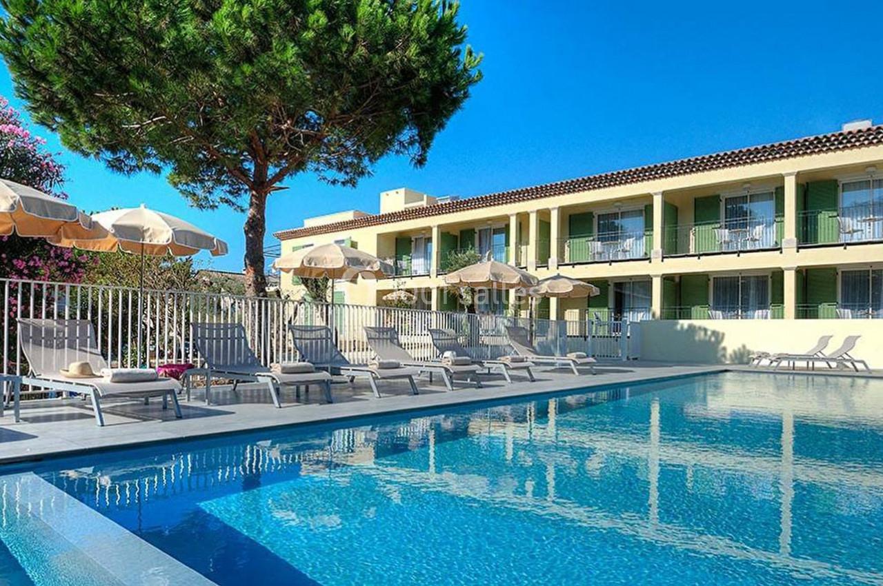 Piscine extérieure entourée de chaises longues et parasols, devant un bâtiment à deux étages avec balcons.