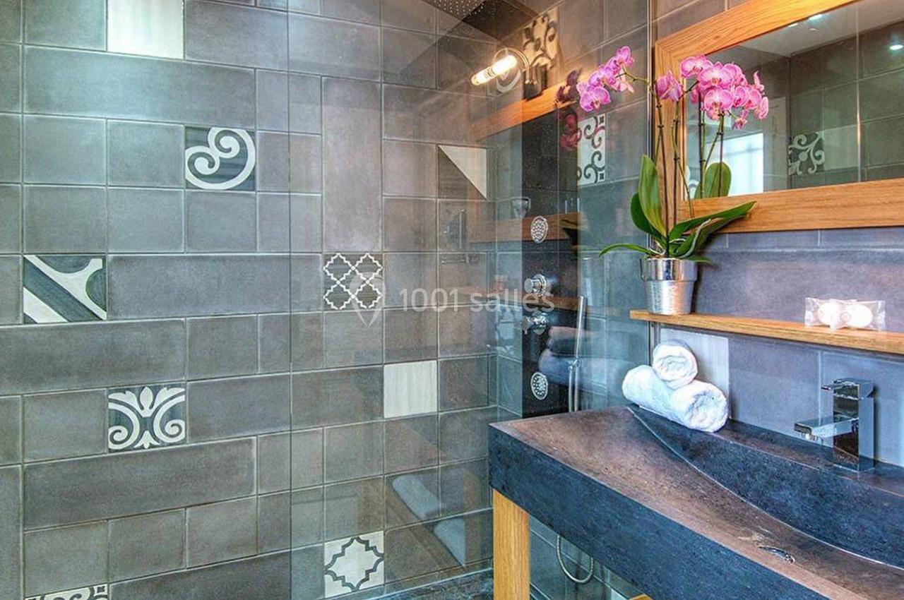 Salle de bain avec douche à carreaux gris ornés de motifs, lavabo en pierre et orchidée rose sur le plan de travail.