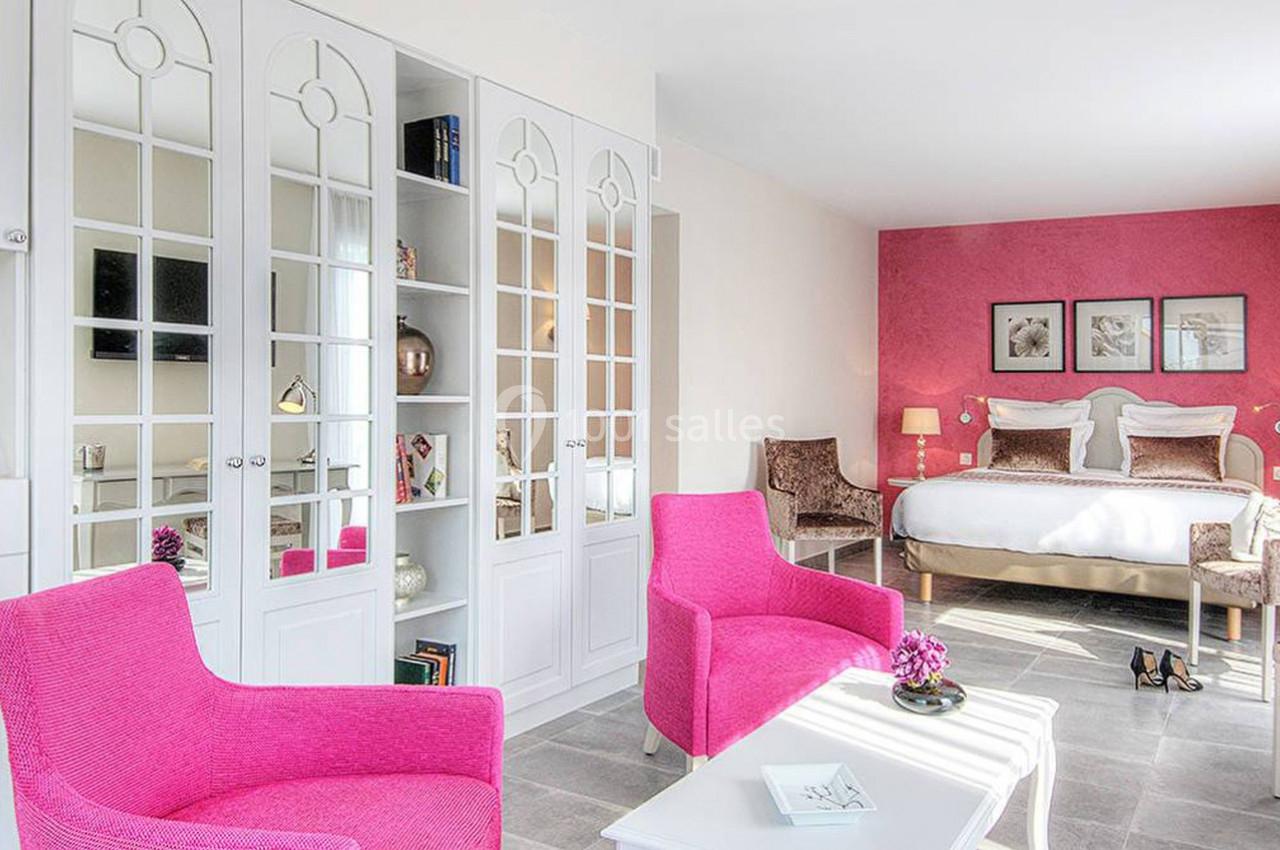 Chambre lumineuse avec un mur rose, deux fauteuils fuchsia, un lit double et des meubles blancs avec miroirs.