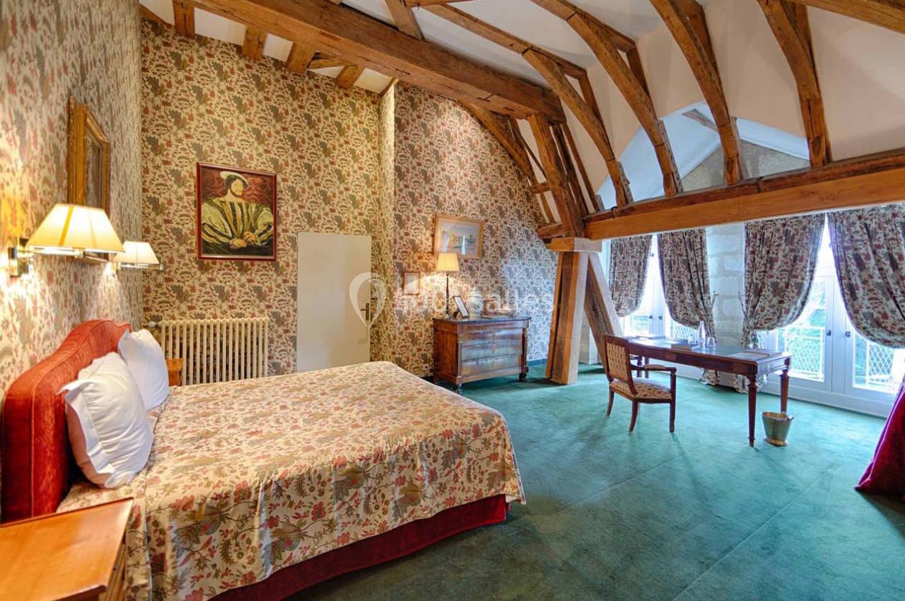 Chambre avec lit double, poutres apparentes, papier peint fleuri, bureau en bois et grandes fenêtres avec rideaux.