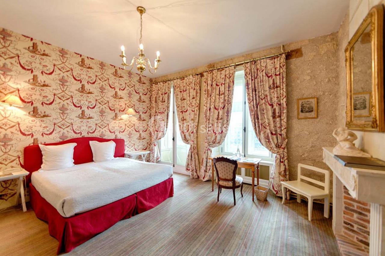 Chambre lumineuse avec lit double, murs décorés de motifs rouges, bureau près de la fenêtre et cheminée en pierre.