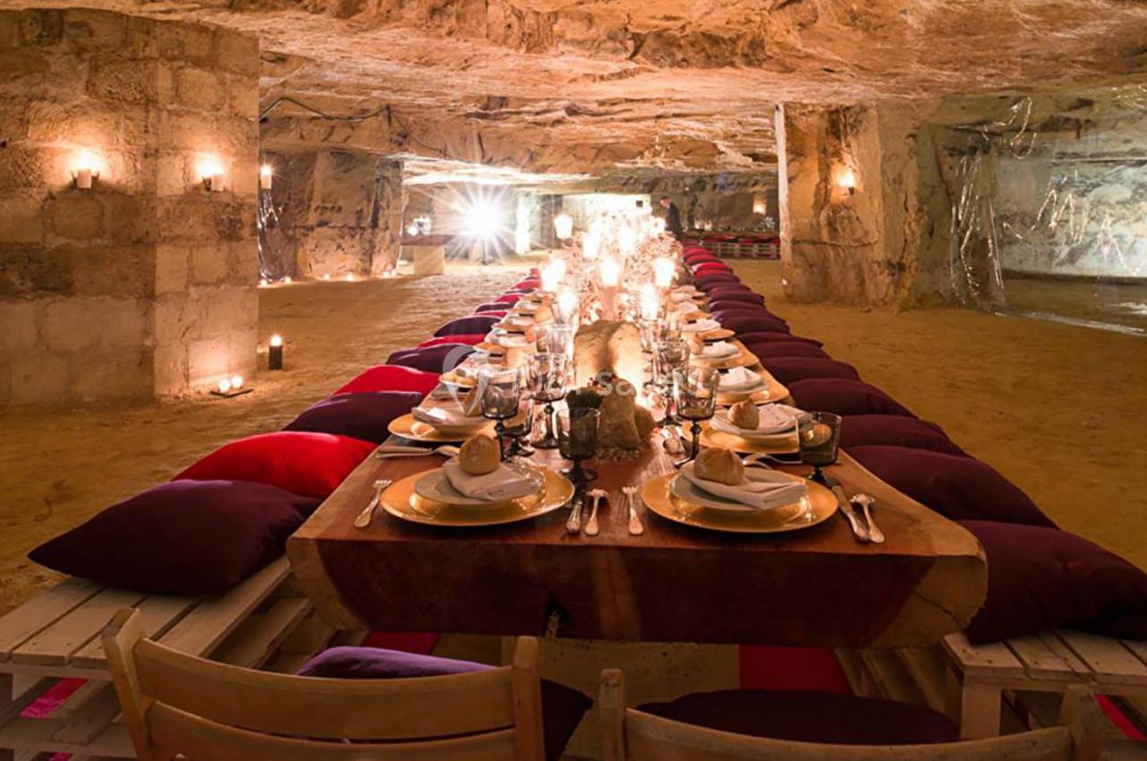 Table dressée pour un dîner dans une grotte éclairée par des bougies, avec coussins colorés et vaisselle élégante.