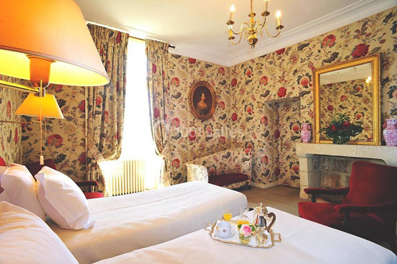 Chambre décorée de motifs floraux avec deux lits, un miroir, des fauteuils rouges et une table avec un service à thé.