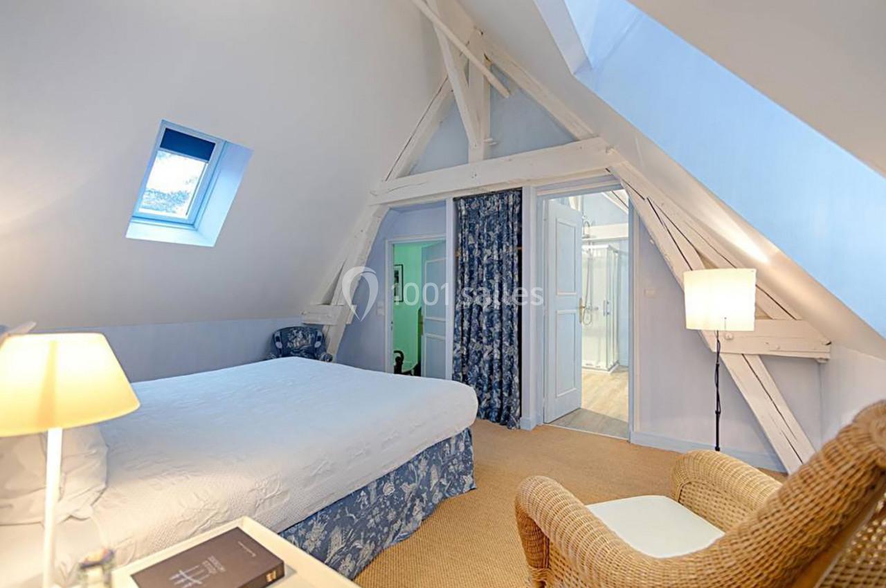 Chambre mansardée lumineuse avec lit double, fauteuil en osier, lampes et accès à une salle de bain adjacente.