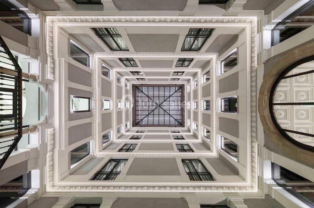 Vue en contre-plongée d'un atrium carré avec des balcons et un plafond vitré laissant entrer la lumière naturelle.