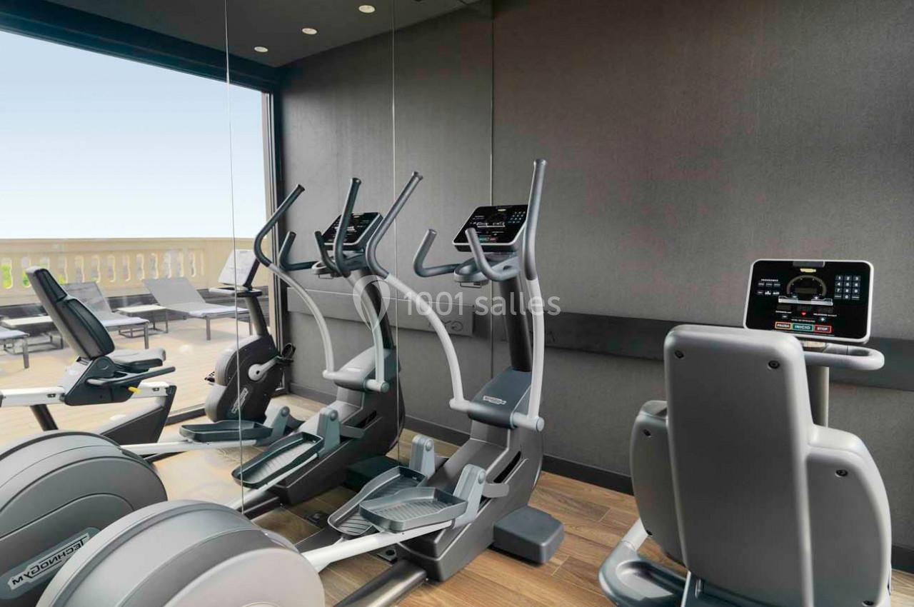 Salle de sport avec vélos elliptiques et tapis de course face à une baie vitrée donnant sur une terrasse extérieure.