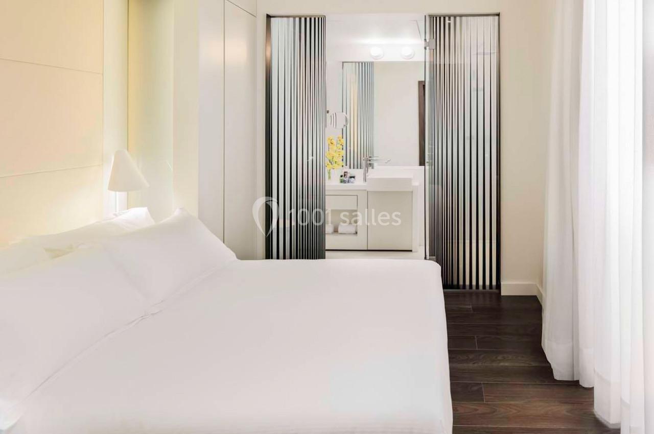 Chambre lumineuse avec lit double, parquet foncé et salle de bain attenante séparée par des panneaux coulissants.