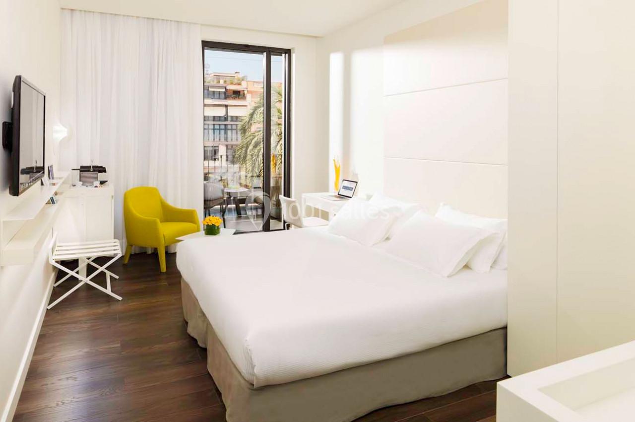 Chambre lumineuse avec lit double, fauteuil jaune, bureau, télévision murale et vue sur des bâtiments depuis une baie vitrée.