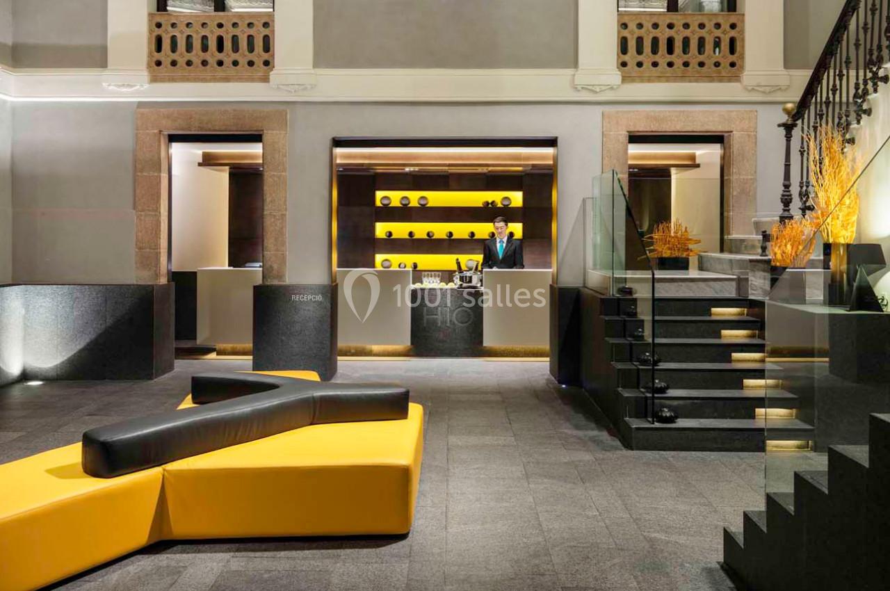 Réception d'un hôtel moderne avec comptoir éclairé, canapé jaune en forme géométrique et escalier en verre.