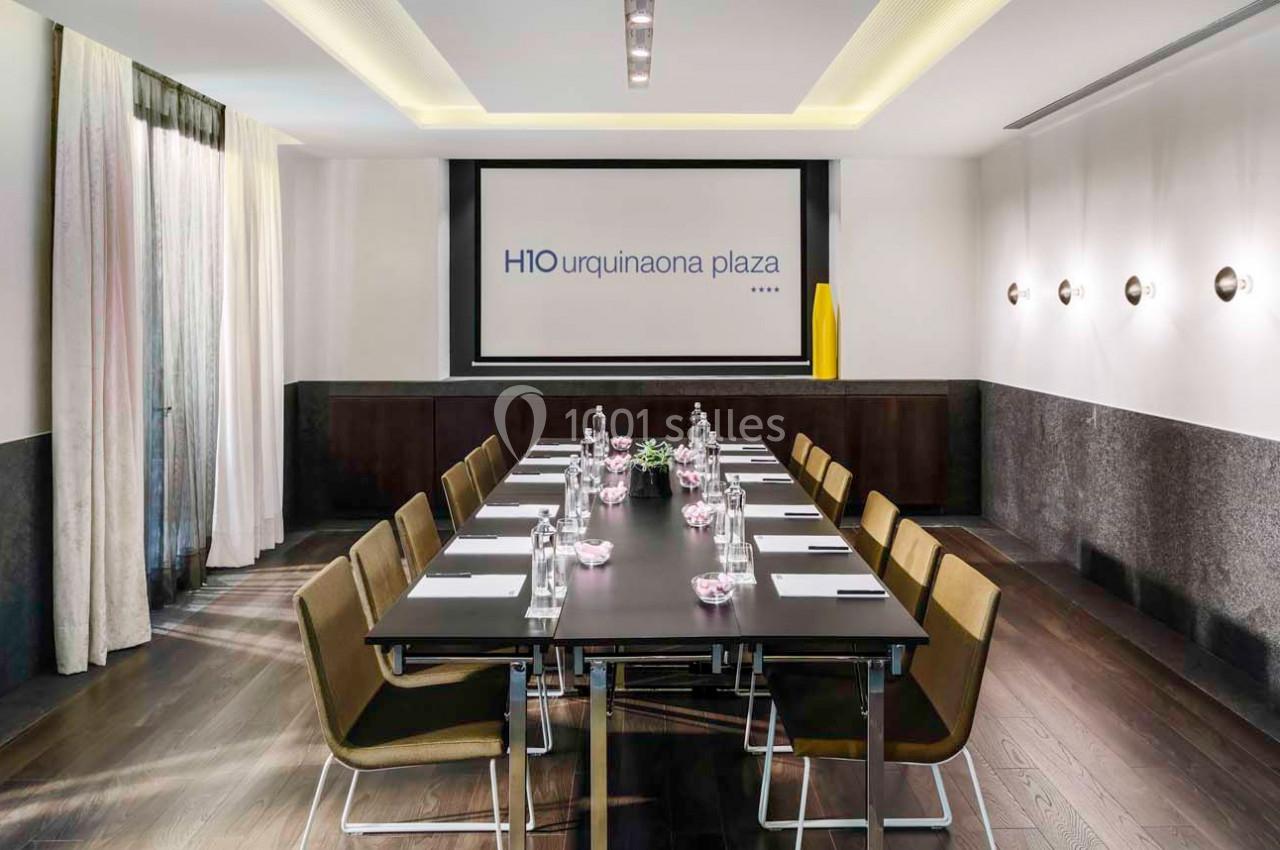 Salle de réunion moderne avec une grande table rectangulaire, chaises, bouteilles d'eau et écran de présentation.