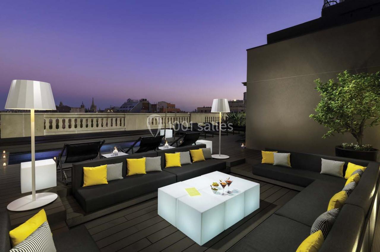 Terrasse moderne avec mobilier sombre, coussins jaunes, table lumineuse et lampes, vue sur un ciel au crépuscule.