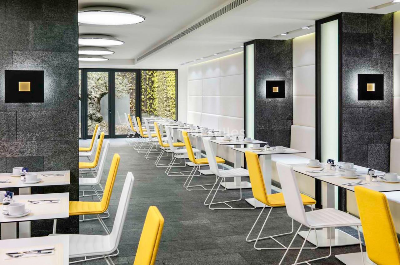 Salle lumineuse avec tables dressées, chaises blanches et jaunes, murs modernes et grandes baies vitrées donnant sur un…