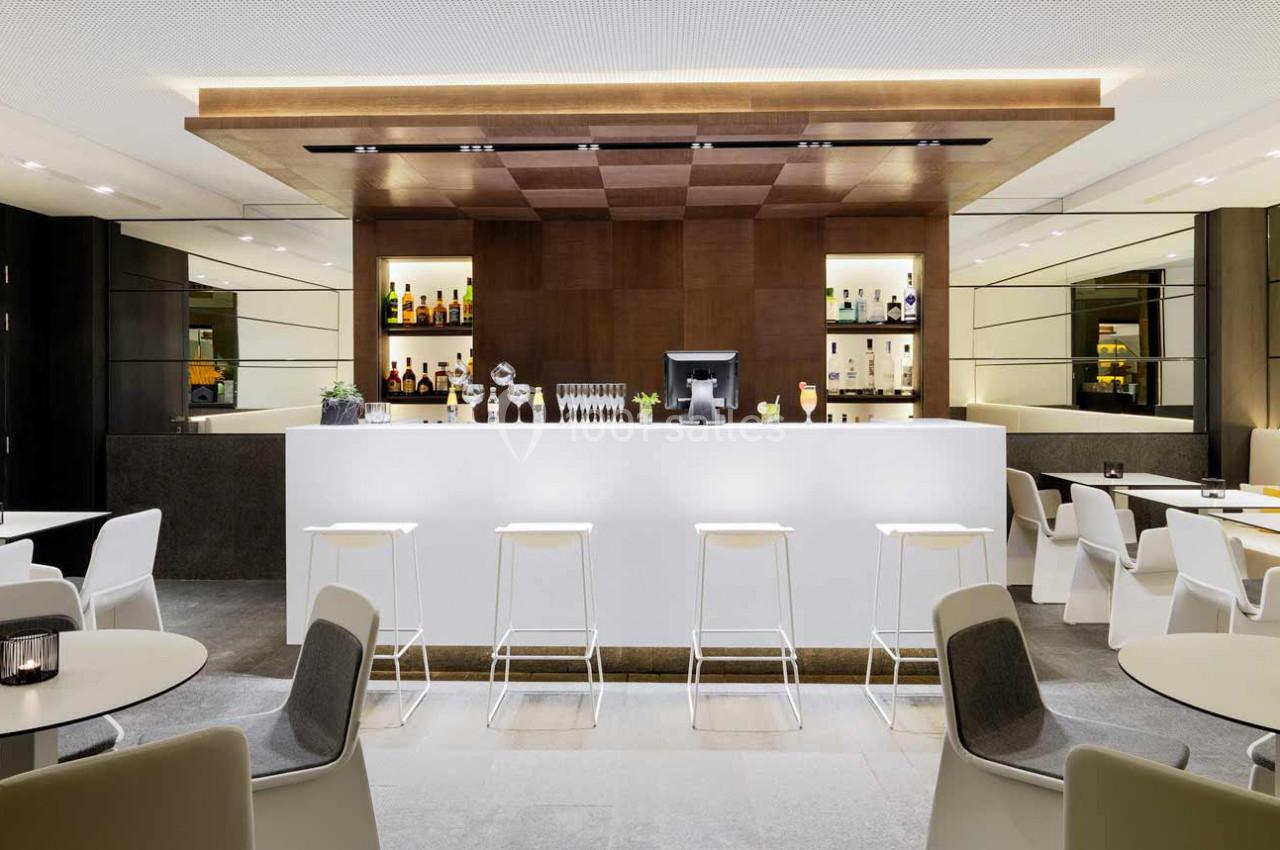 Bar moderne avec comptoir blanc, tabourets alignés, étagères éclairées contenant des bouteilles et verres en arrière-plan.