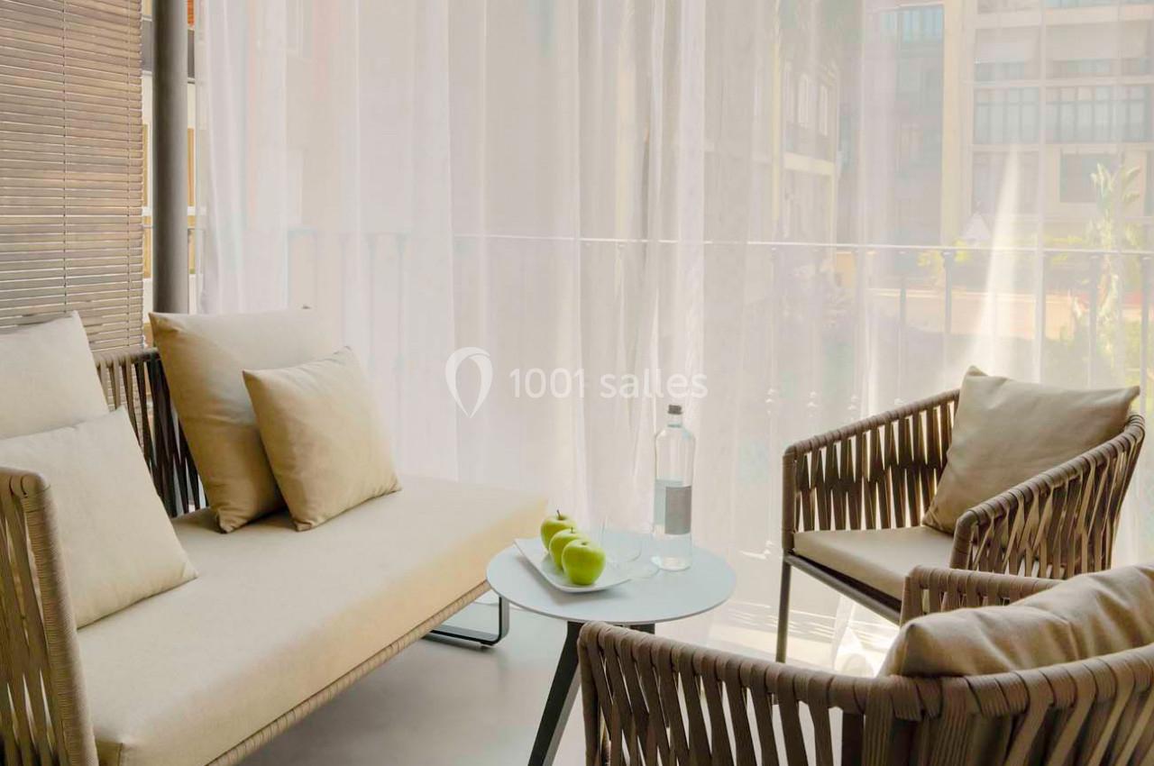 Salon extérieur avec canapé, fauteuils en rotin, table basse avec bouteille d'eau et pommes, devant un rideau translucide.