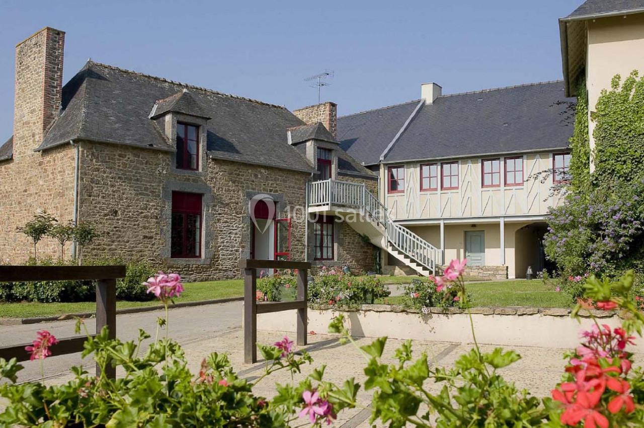 Maisons traditionnelles en pierre avec volets rouges et colombages, entourées de fleurs et d'une cour pavée.