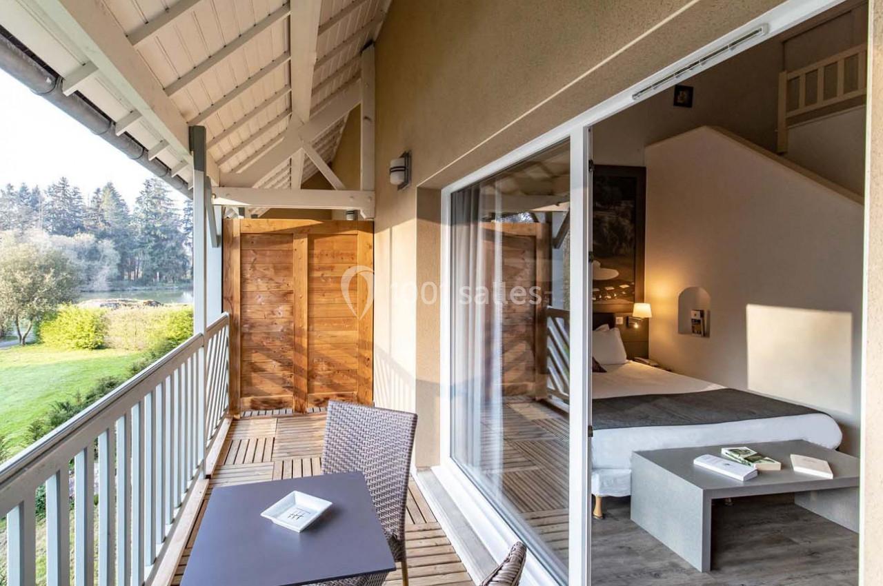 Vue d'une chambre d'hôtel avec balcon, table et chaises, donnant sur un paysage verdoyant et un plan d'eau.