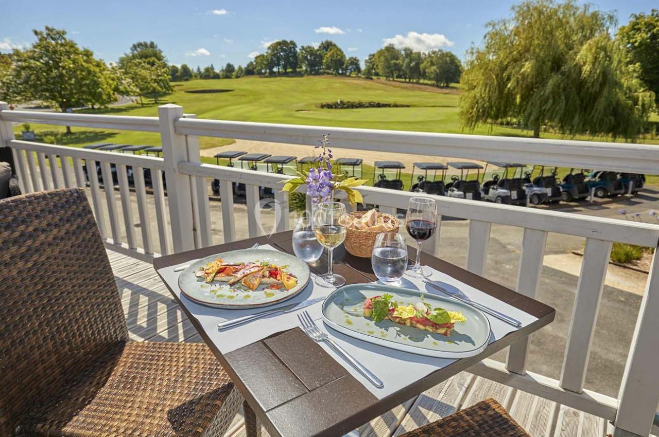 Table en terrasse avec deux assiettes de plats raffinés, verres de vin et vue sur un terrain de golf ensoleillé.
