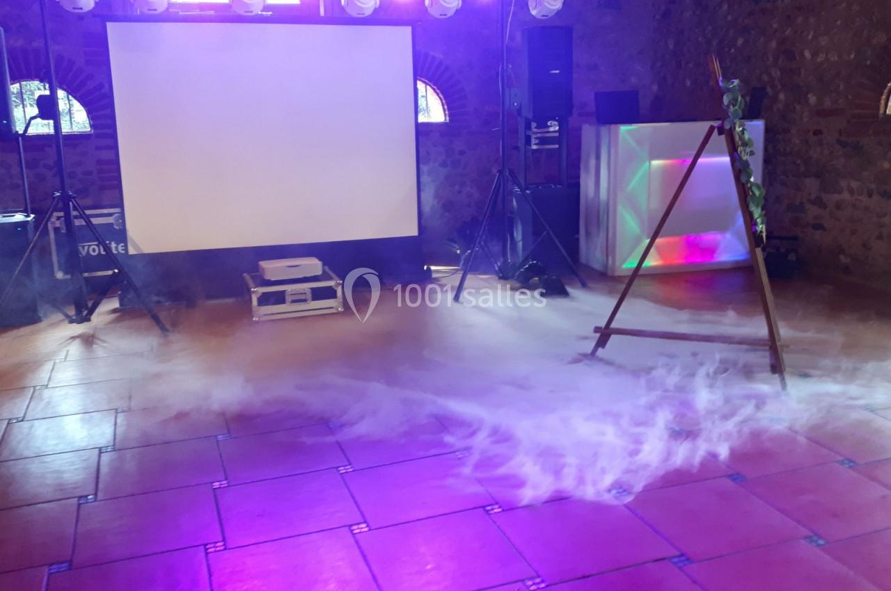 Salle équipée pour un événement avec écran de projection, éclairage coloré et sol recouvert de fumée légère.