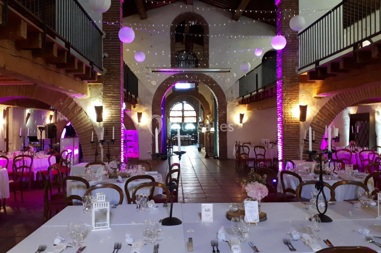 Salle de réception décorée pour un événement avec tables dressées, guirlandes lumineuses et éclairage violet.
