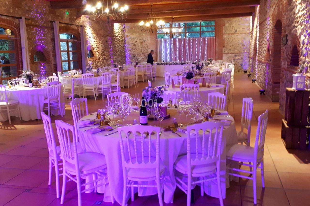 Salle de réception décorée pour un événement, avec tables rondes dressées, chaises blanches et éclairage tamisé.