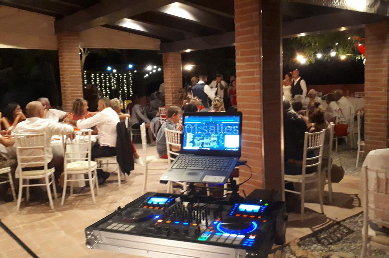 Table de mixage et ordinateur installés sous une pergola, avec des invités assis à des tables lors d'une soirée en extérieur.