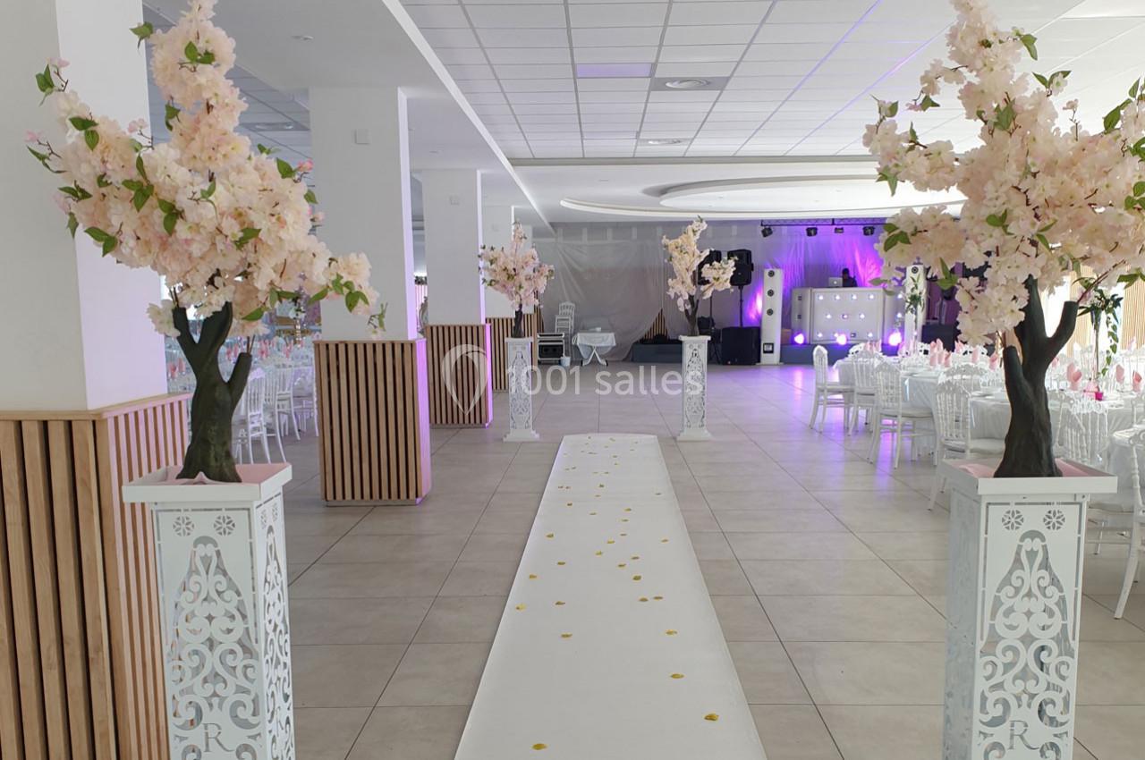 Salle de réception décorée avec des arbres fleuris, une allée blanche parsemée de pétales et des tables dressées en arrière…