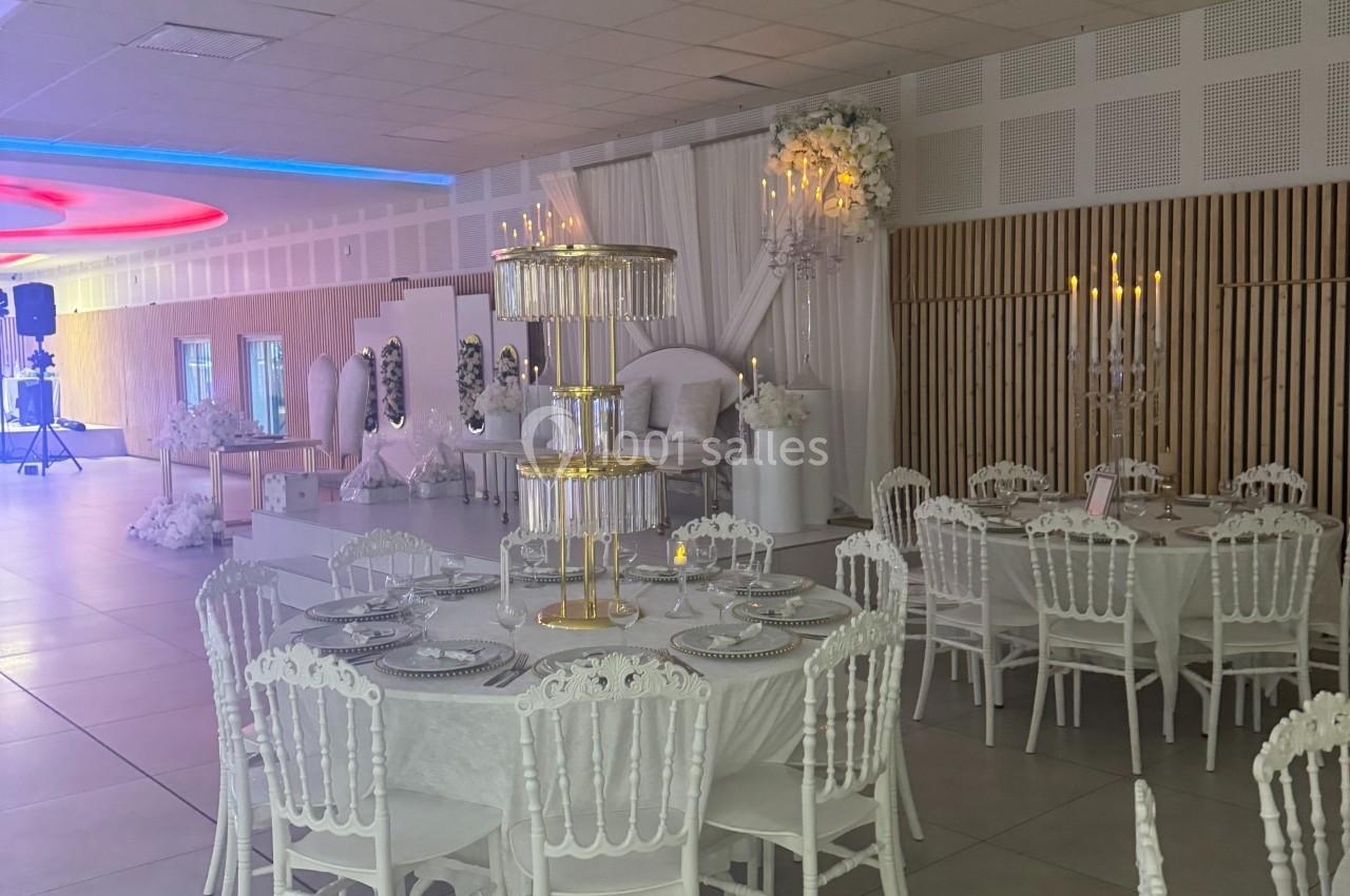 Salle de réception décorée en blanc avec des tables rondes, chaises assorties et un présentoir central pour desserts.