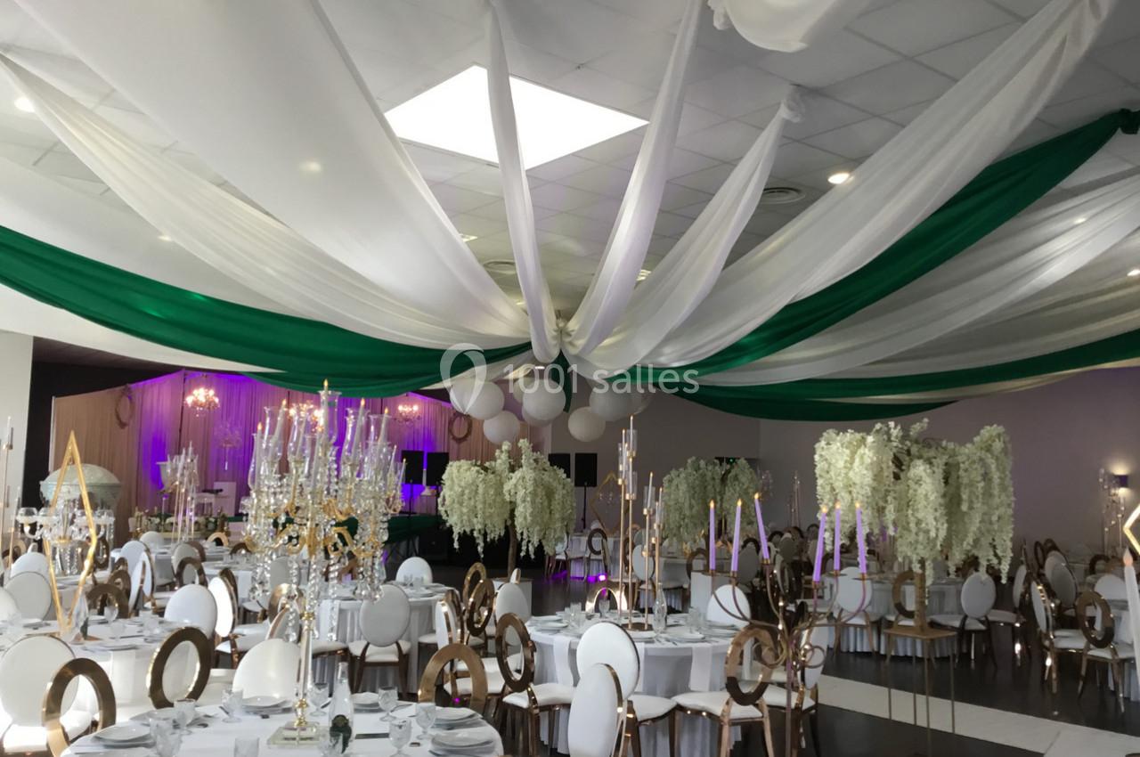 Salle de réception décorée avec des tables rondes, chaises élégantes, drapés verts et blancs au plafond, et fleurs…