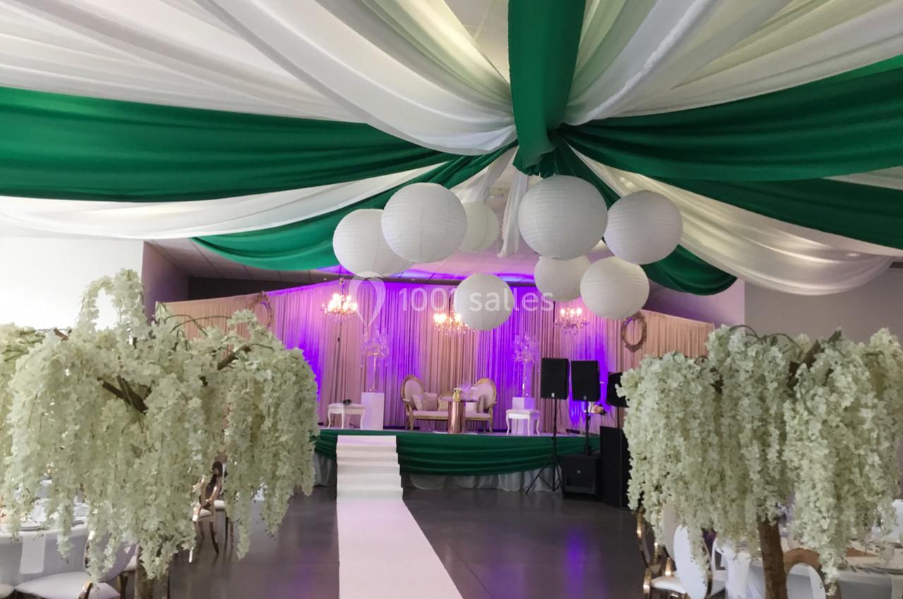 Salle de réception décorée avec des drapés verts et blancs, des lanternes suspendues et des arbres artificiels fleuris.