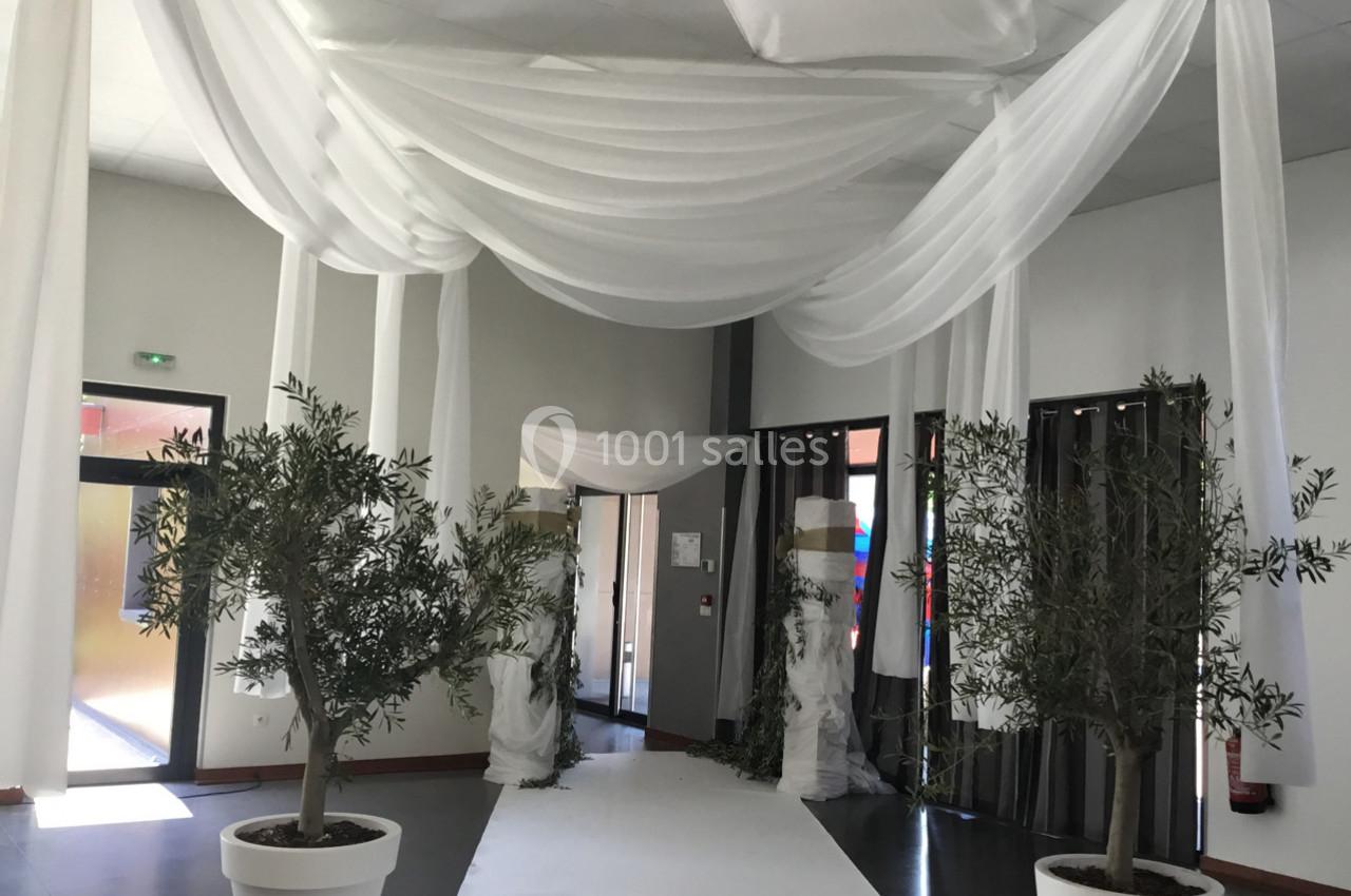 Salle décorée pour un événement avec drapés blancs suspendus, tapis blanc au sol et plantes en pots.