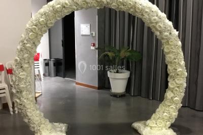 Arc décoratif en fleurs blanches placé dans une salle avec des rideaux gris et un sol en béton.
