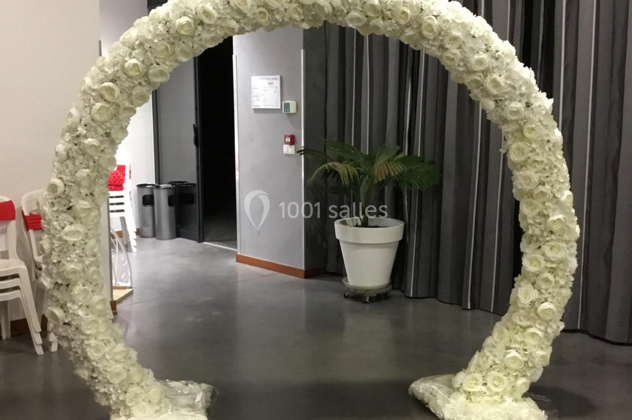 Arc décoratif en fleurs blanches placé dans une salle avec des rideaux gris et un sol en béton.