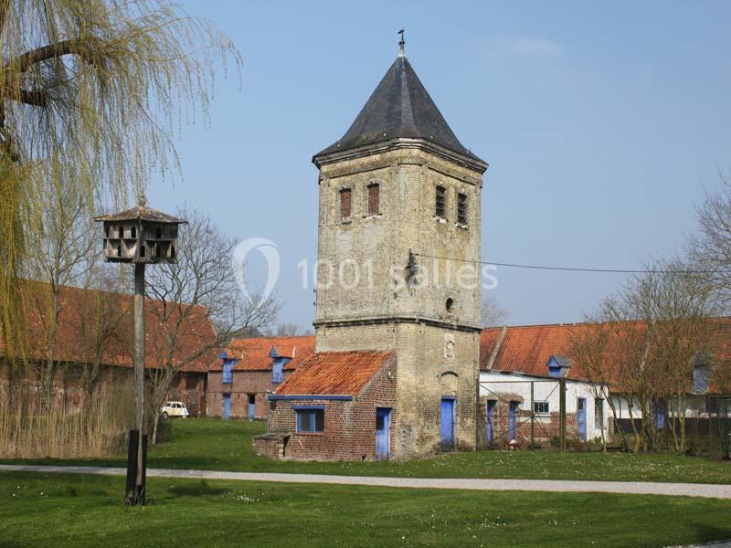 Tour en briques avec toit en ardoise, entourée de bâtiments de ferme et d'arbres, sous un ciel dégagé.