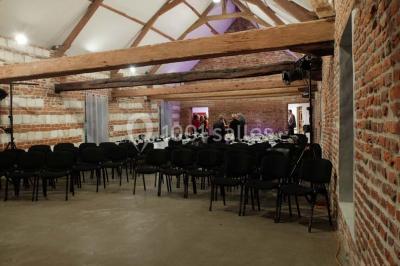 Façade d'une grange en briques avec porte en bois rouge, toit en tuiles et piles de bois empilées sur les côtés.