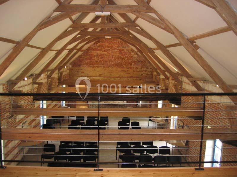 Salle avec charpente en bois apparente, murs en briques et rangées de chaises noires disposées pour un événement.