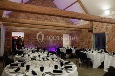 Façade d'une grange en briques avec porte en bois rouge, toit en tuiles et piles de bois empilées sur les côtés.