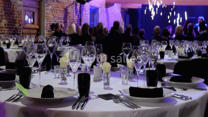 Salle de réception avec tables dressées, nappes blanches, verres, couverts et décorations florales, éclairage tamisé.