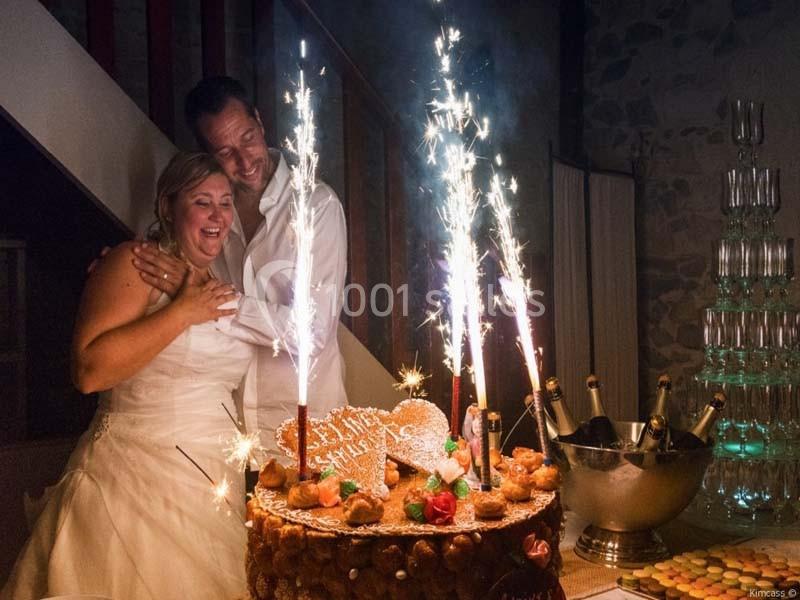 Un couple en tenue de mariage sourit devant une pièce montée décorée de cierges étincelants.