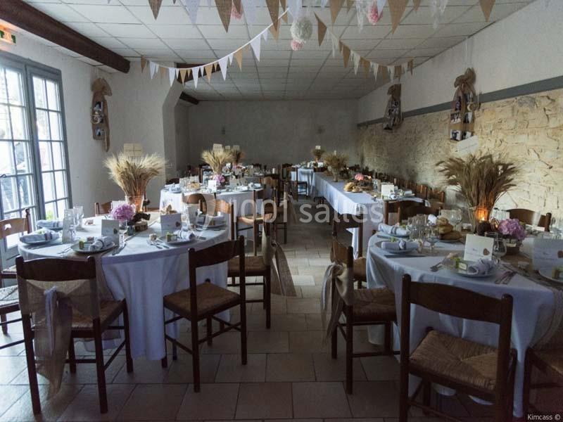 Salle décorée pour un événement, avec tables rondes dressées, nappes blanches, bouquets de fleurs et épis de blé.