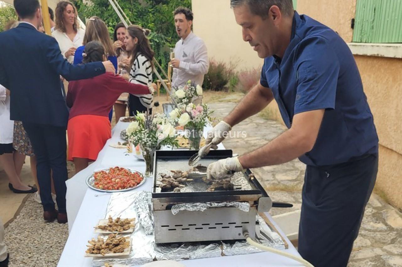 Un cuisinier prépare des grillades sur une plancha lors d'un événement en extérieur avec des invités en arrière-plan.