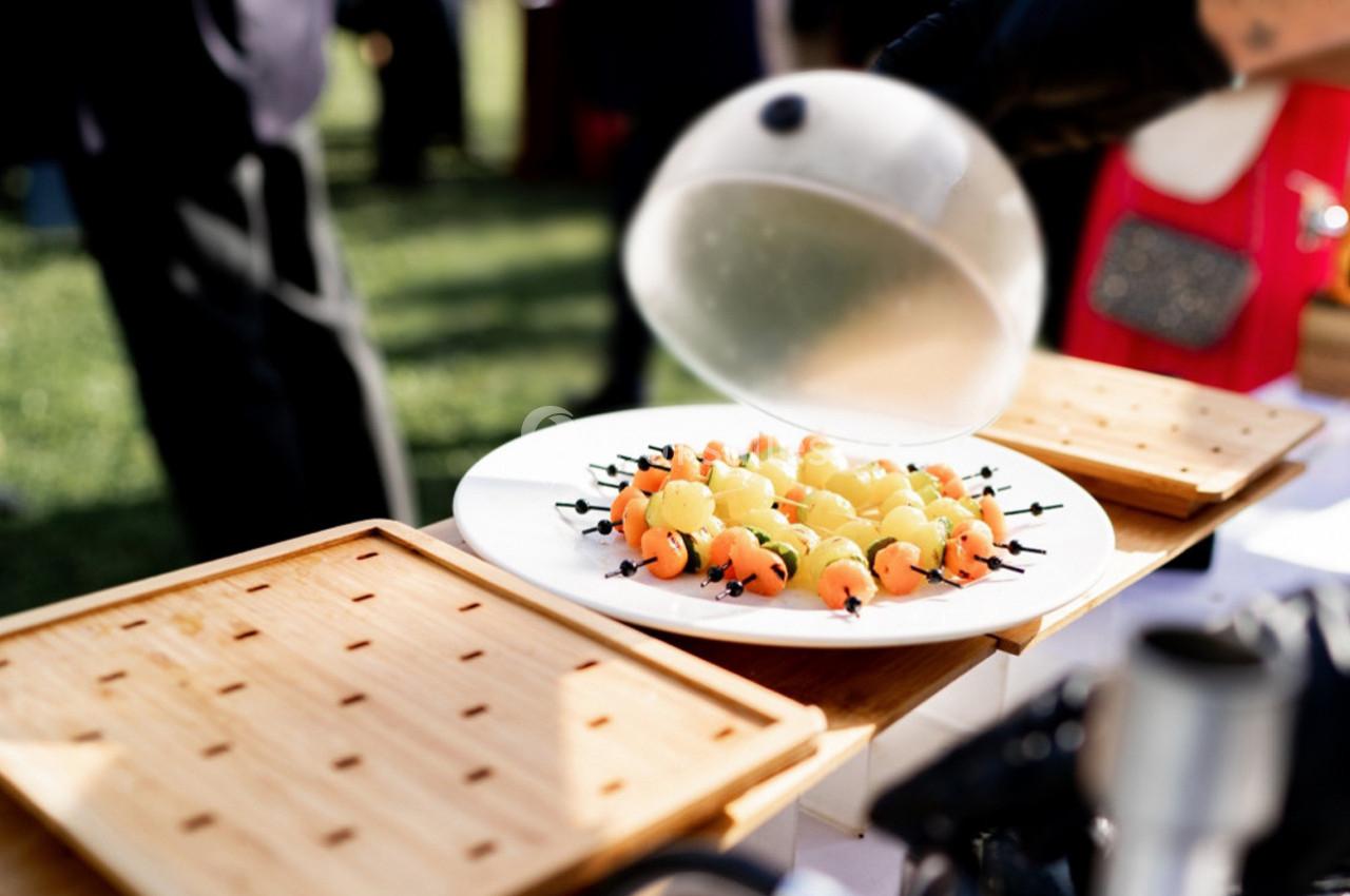 Assiette de brochettes de fruits colorés présentée sous une cloche transparente lors d'un événement en extérieur.