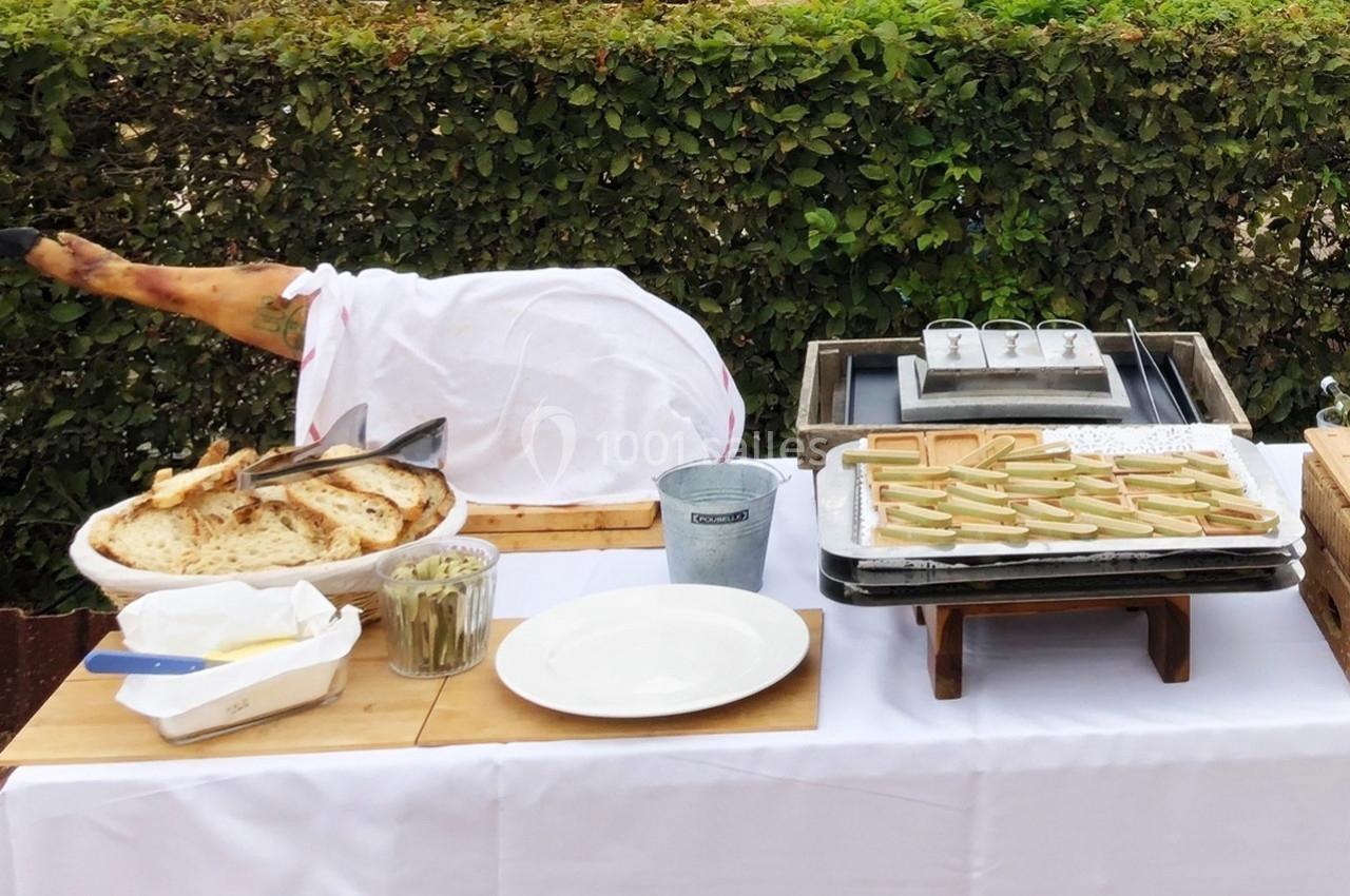 Table de buffet en extérieur avec jambon cru, pain, beurre, olives et plateaux de tapas devant une haie verte.