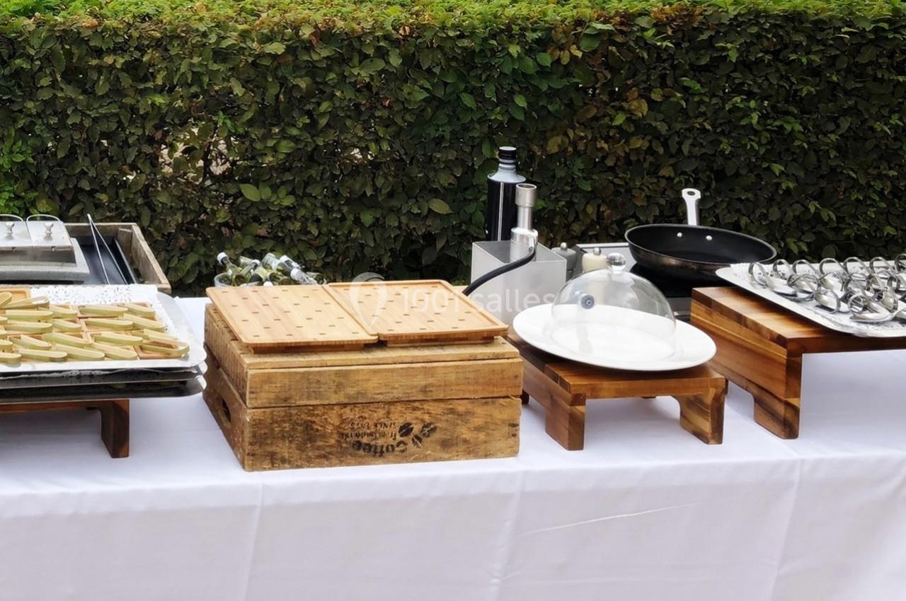 Table de buffet en extérieur avec plats variés, ustensiles et présentoirs en bois sur une nappe blanche.