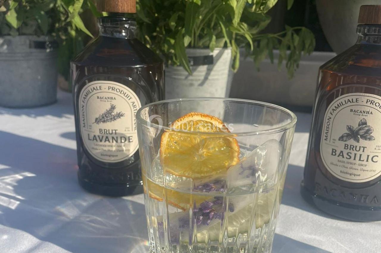Verre de cocktail garni d'une tranche d'orange séchée et de fleurs, entouré de bouteilles d'aromates sur une table blanche.