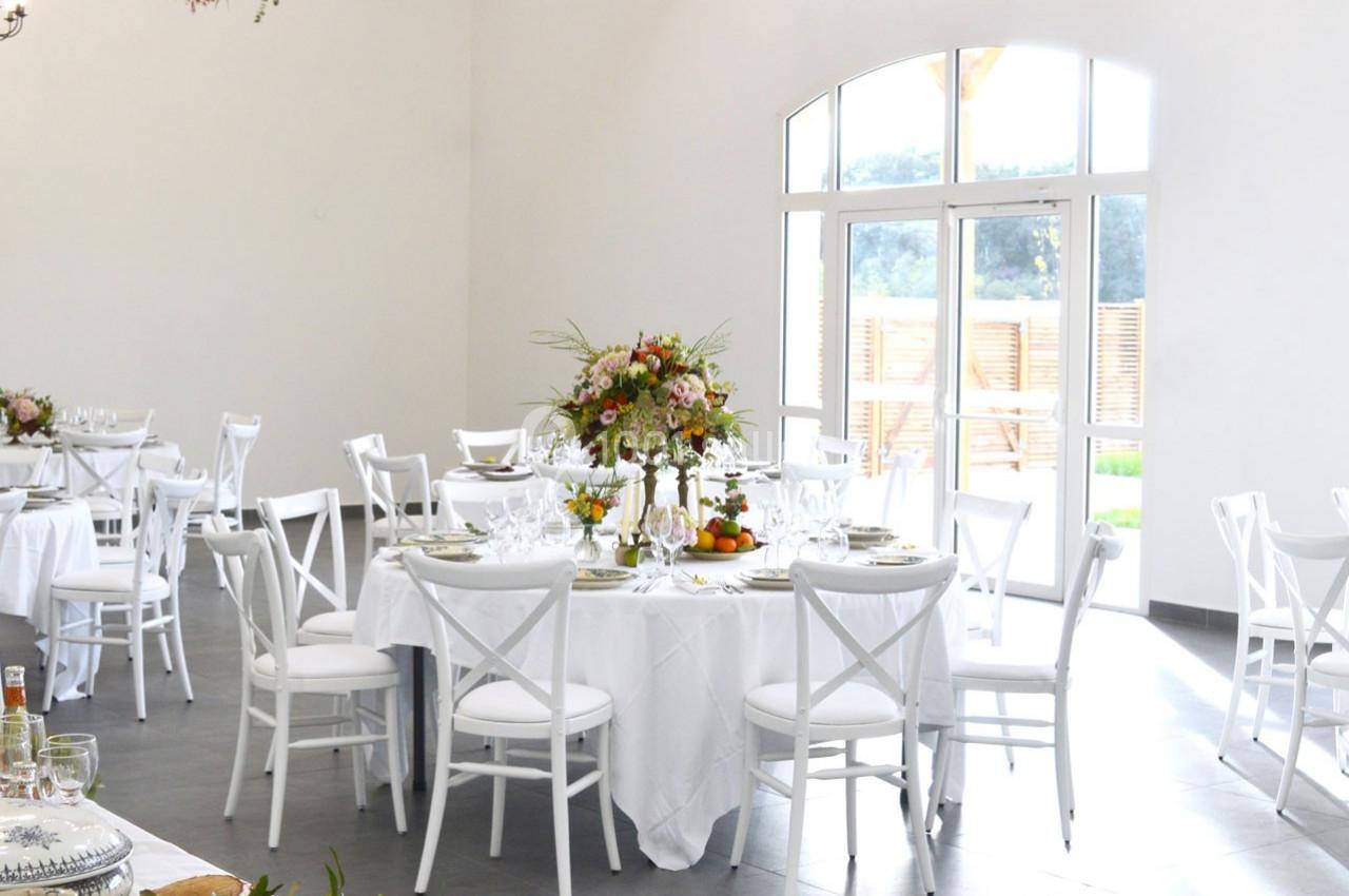 Salle lumineuse avec tables rondes dressées, nappes blanches, chaises blanches et centre de table floral coloré.