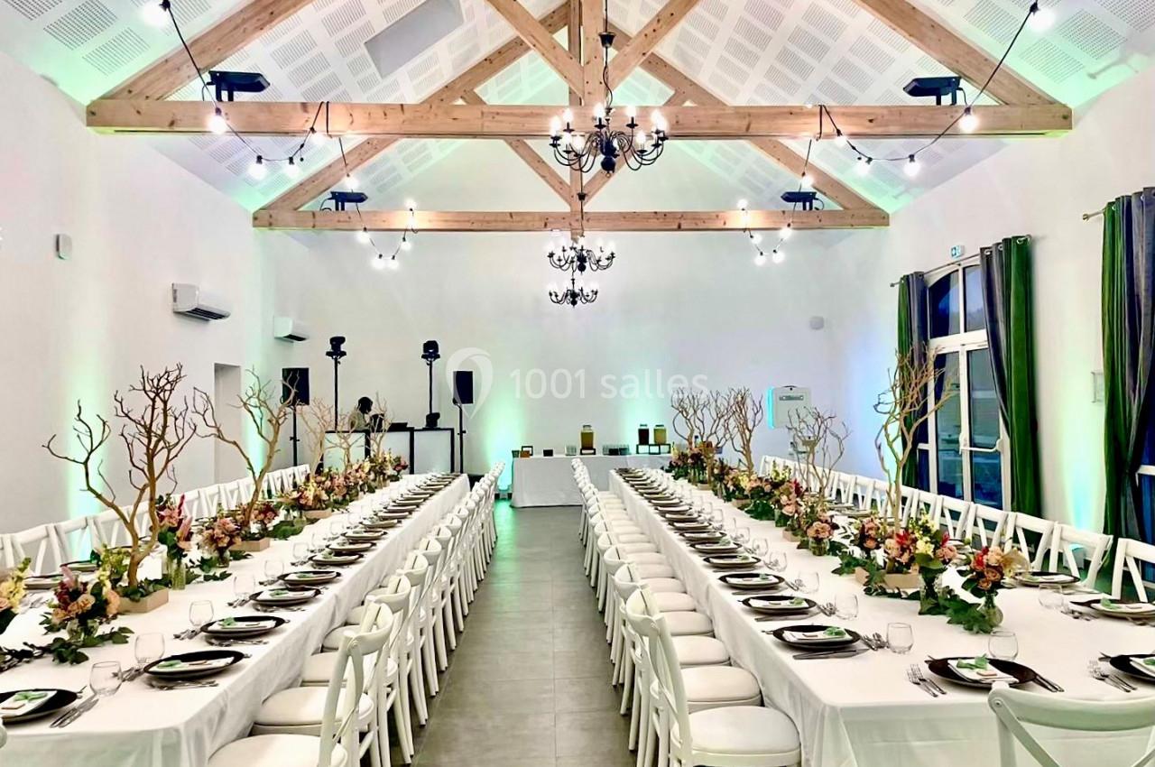 Salle de réception lumineuse avec deux longues tables décorées de fleurs et branches, sous un plafond à poutres apparentes.